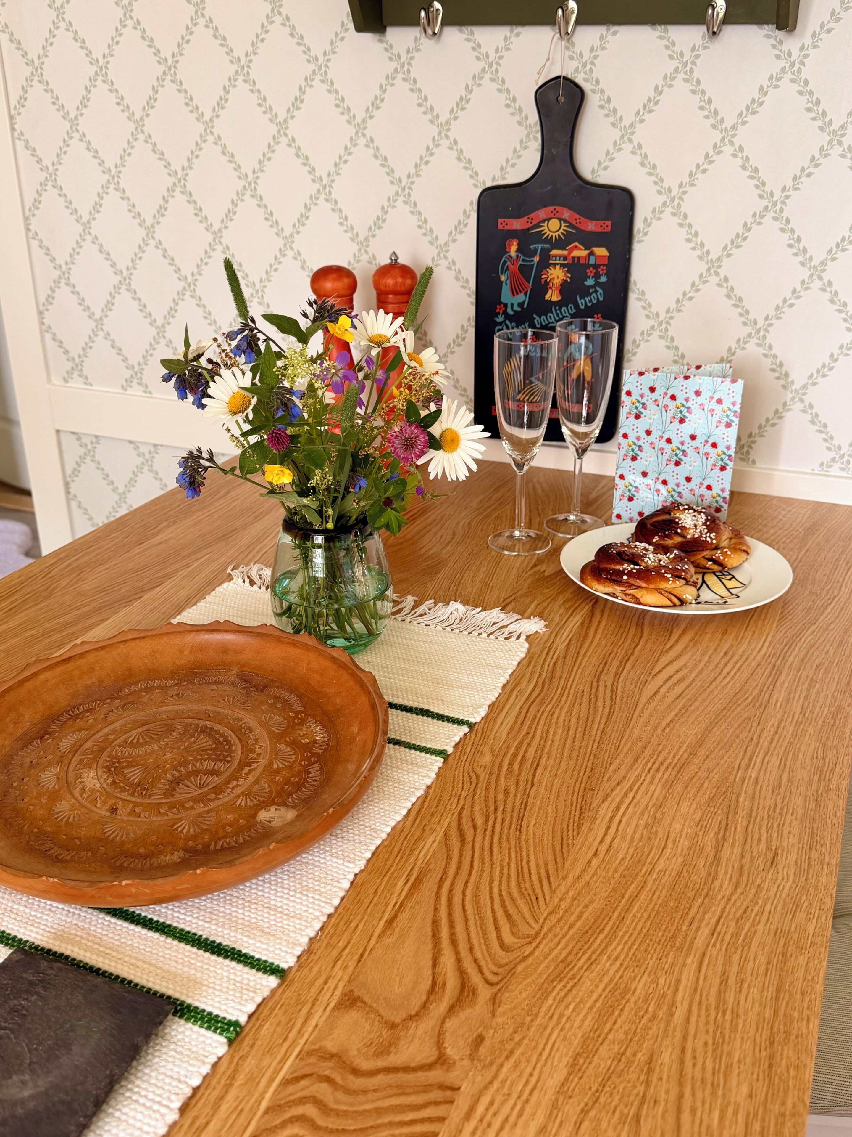 Een houten tafel met een geweven placemat, een decoratieve schaal, een vaas met bloemen, twee wijnglazen, een bord met gebak en een lijst met een foto.