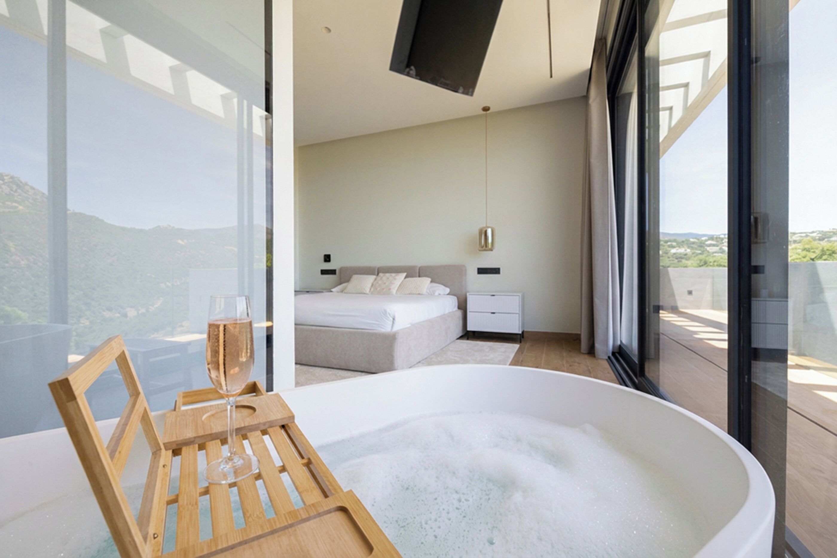 Un dormitorio moderno con una bañera grande, una mesita de noche con una copa de champán y vistas al exterior.