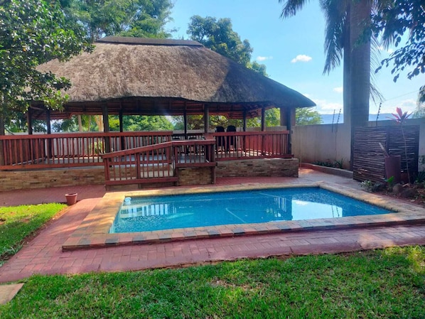 A pool area with a thatched-roof structure, a wooden deck, and a stone wall.