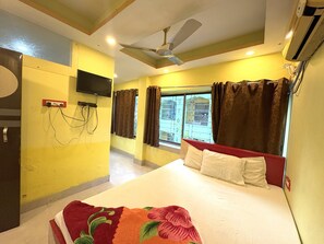 A hotel room with a bed, a ceiling fan, an air conditioning unit, a TV mounted on the wall, and a window with curtains. - Inn Swastika on Forest Bungalow Rd Digha (Digha)