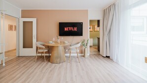 Ein modernes Wohnzimmer mit einem runden Holztisch, zwei Stühlen, einem flachbildfernseher an der Wand und einem großen Fenster mit transparenten Vorhängen.