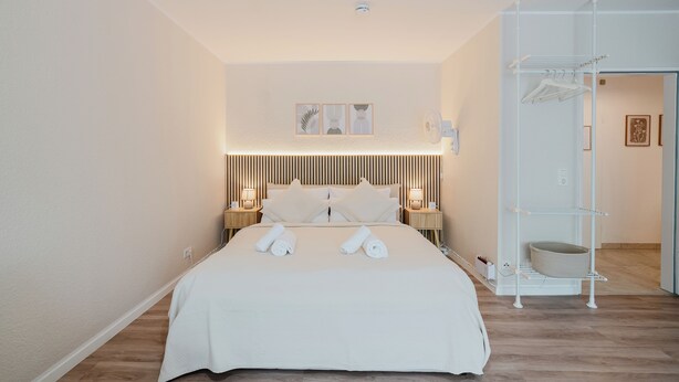 Ein ordentlich bezogenes Bett mit Handtüchern, einer Nachttischlampe, einem hängenden Regal und einer Wandablage.