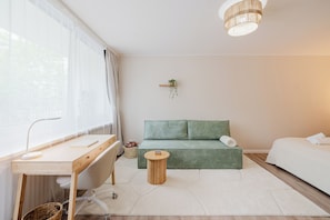 Ein minimalistisch eingerichteter Raum mit einem Holztisch, einem grünen Sofa, einem Bett, einem kleinen runden Tisch und einer hängenden Pflanze.