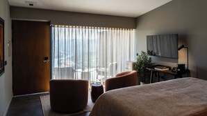 Habitación de hotel con cama, dos sillones, un escritorio con lámpara, una planta en maceta y vistas al exterior a través de cortinas translúcidas.