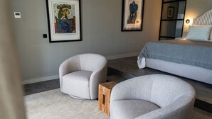Un dormitorio moderno con una cama, dos sillones, un taburete de madera y dos cuadros en la pared.