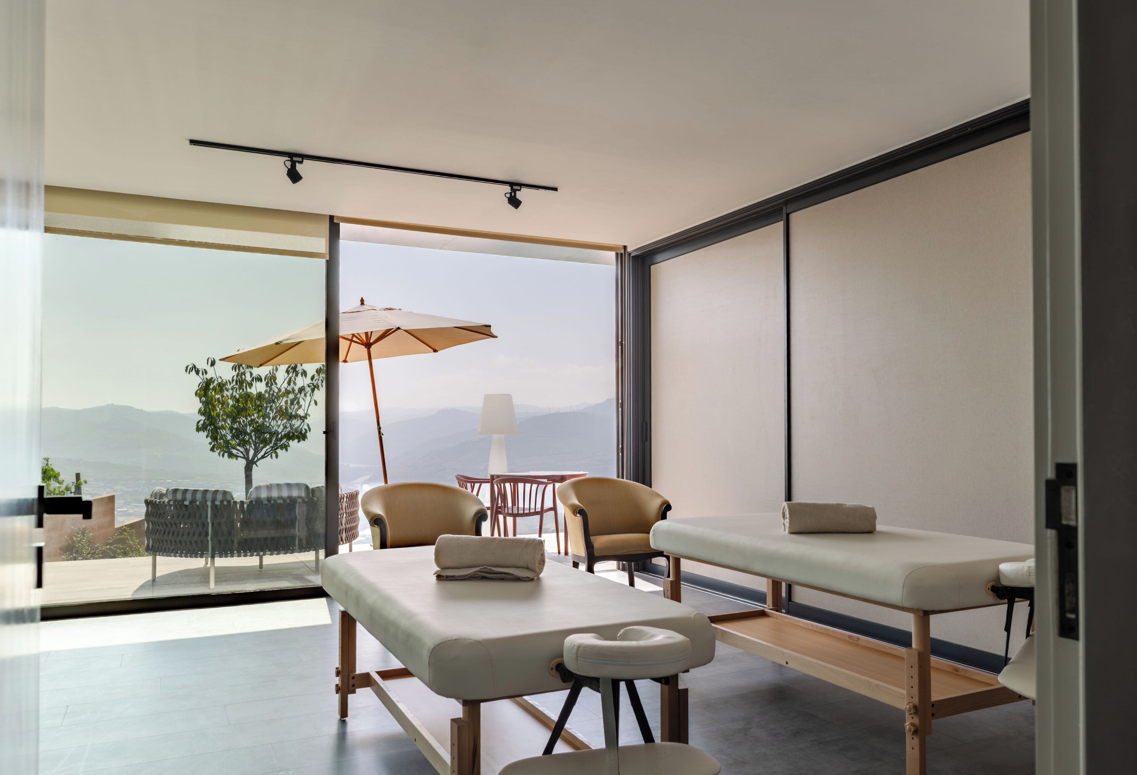 A modern spa room with a massage table, two chairs, a small table with a lamp, and a large window offering a mountain view.