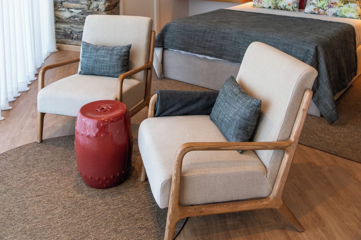Een moderne slaapkamer met een accentmuur van steen, een bed met bloemenkussens en een zithoek met twee stoelen en een rode kruk.