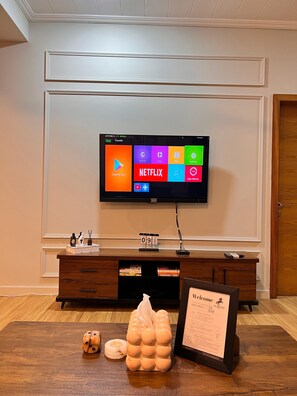 Ein Hotelzimmer mit einem wandmontierten Flachbildfernseher, einem Holzkonsolentisch mit Schubladen, einer kleinen Uhr und einem Willkommensschild.