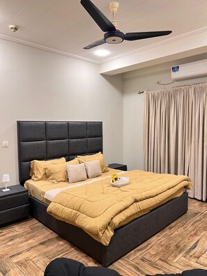 A hotel room with a bed, a nightstand, a ceiling fan, an air conditioning unit, and a curtain.