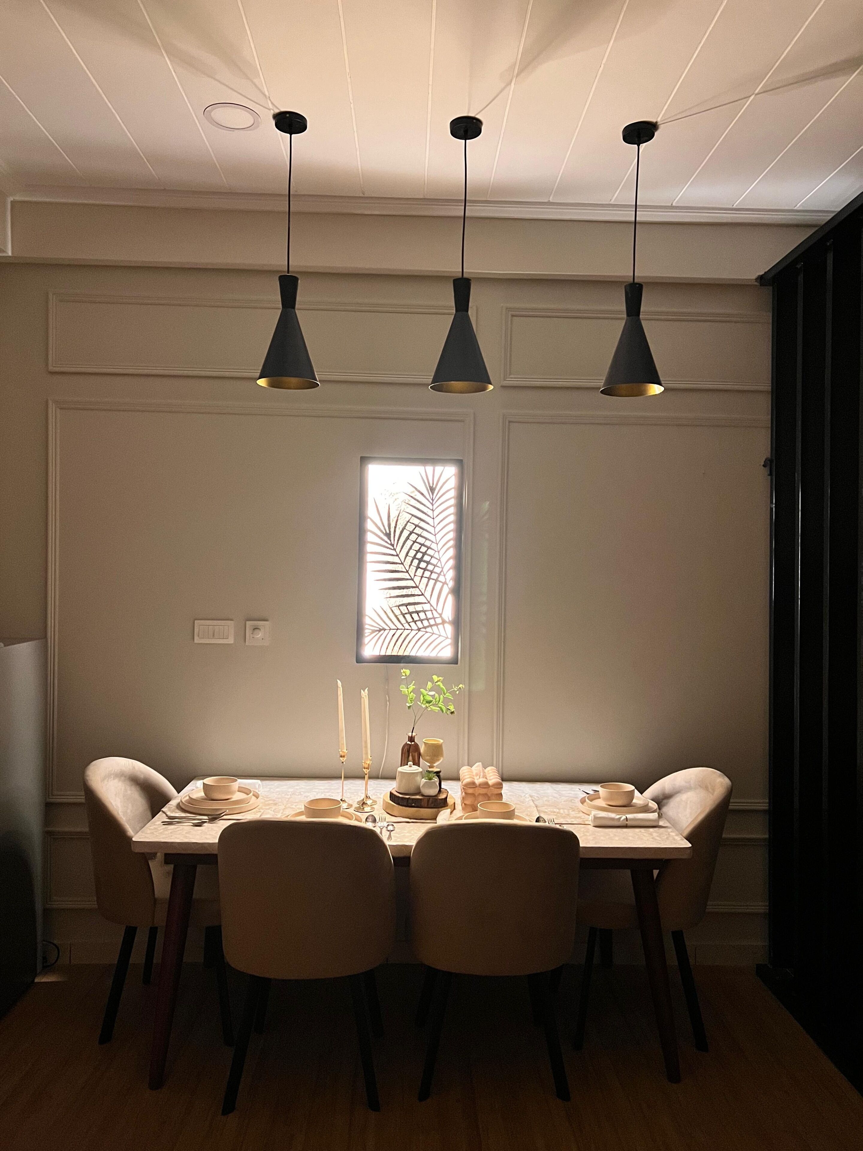 A dining area with a table set for four, pendant lights, a window with a leaf pattern, and a plant on the table.