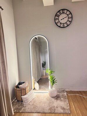 A room with a large mirror, a clock, a potted plant, a small wicker basket, and a textured rug.