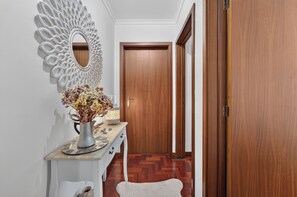 A hallway with a wooden door, a white console table with a vase of flowers, and a decorative mirror on the wall. - Sea Breeze Pilar (Funchal)