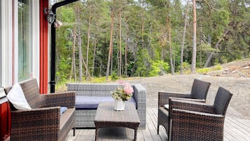 Uma área externa com assento em vime, mobiliário e uma mesa, cercada por um deck de madeira e um fundo de floresta.