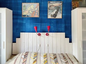 A bedroom with a blue textured wall, two framed artworks, a white headboard, and a quilted bedspread.