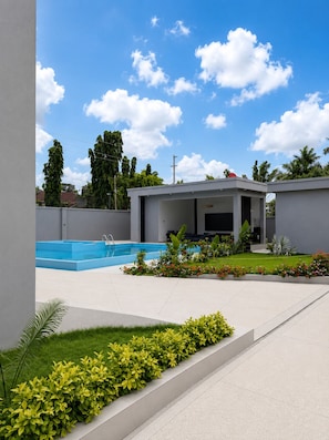 A modern outdoor pool area with a clear blue sky, a covered seating area, and well-maintained greenery.