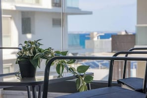 A balcony with a potted plant, a glass table, and a view of buildings and the sea. - Mackenzie Signature Studio Suite in Larnaca (Larnaka)