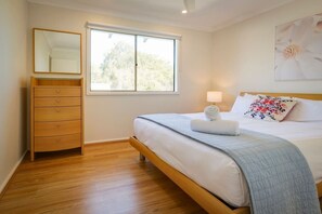 Een slaapkamer met een houten dressoir, een bed met een blauwe deken, een raam en een wandlamp.