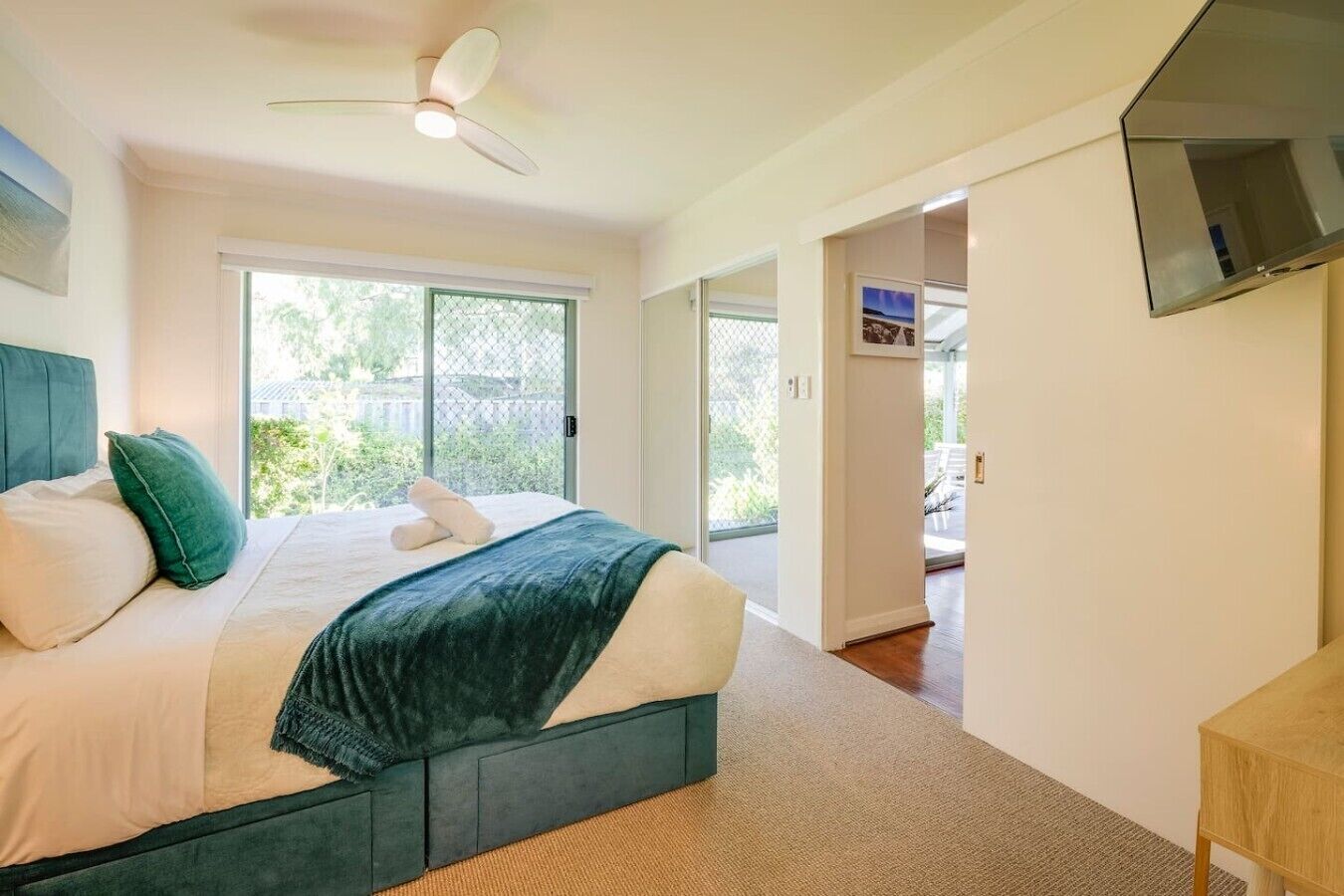 A bedroom with a bed, a ceiling fan, a TV mounted on the wall, and a sliding glass door leading to an outdoor area.