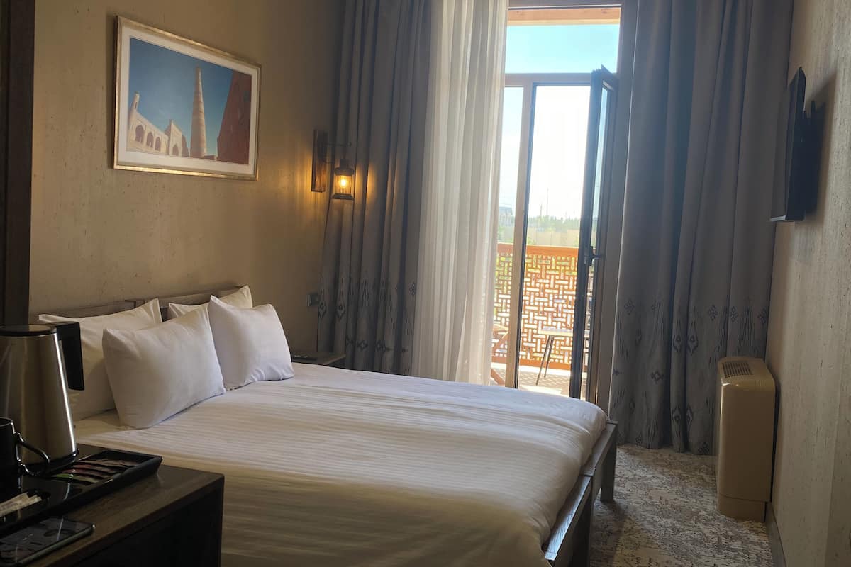A hotel room with a bed, a desk, a painting, a window with curtains, and a ceiling light.
