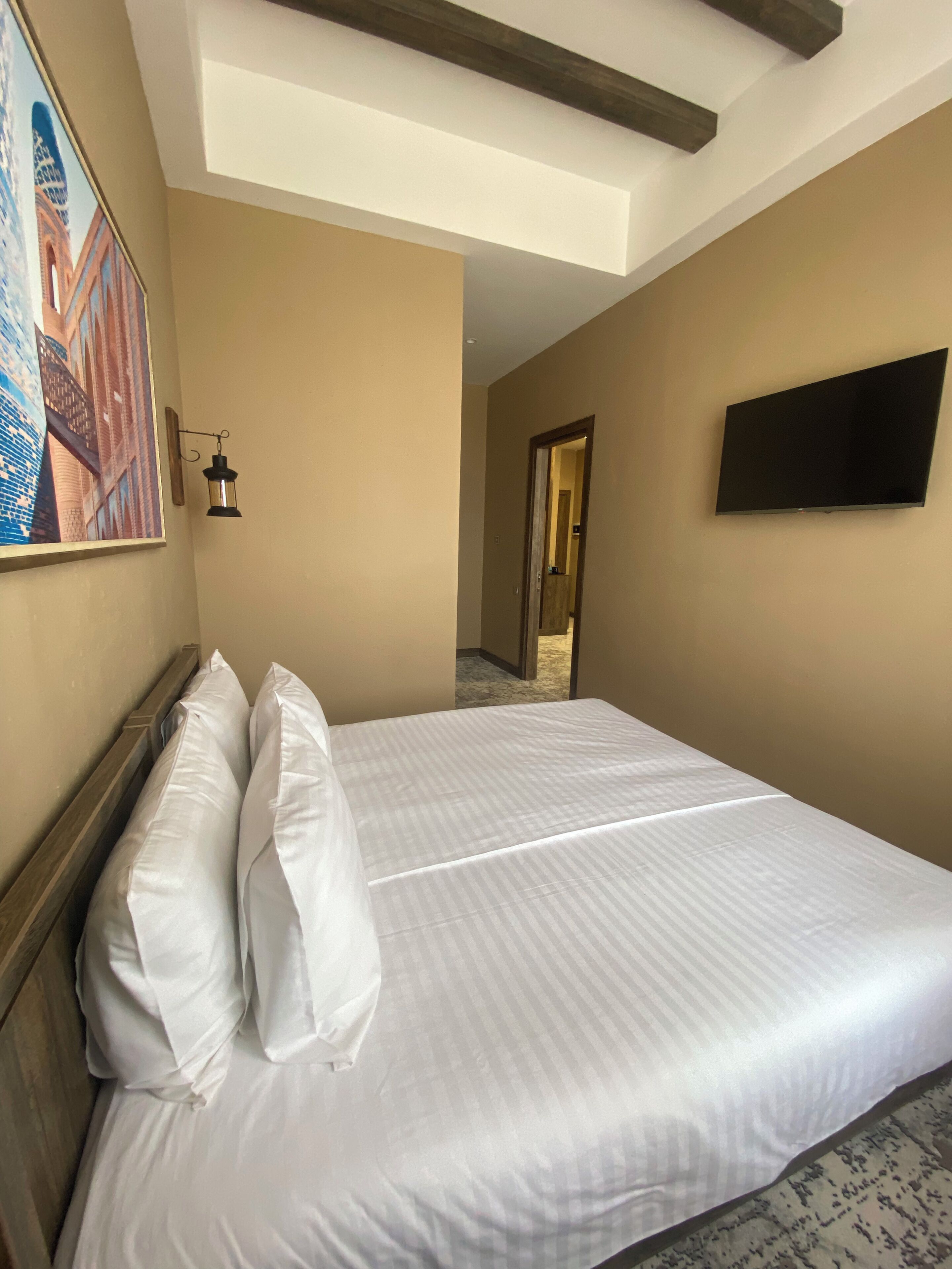 A hotel room with a bed, a painting on the wall, a mounted TV, and a hallway leading to another room.