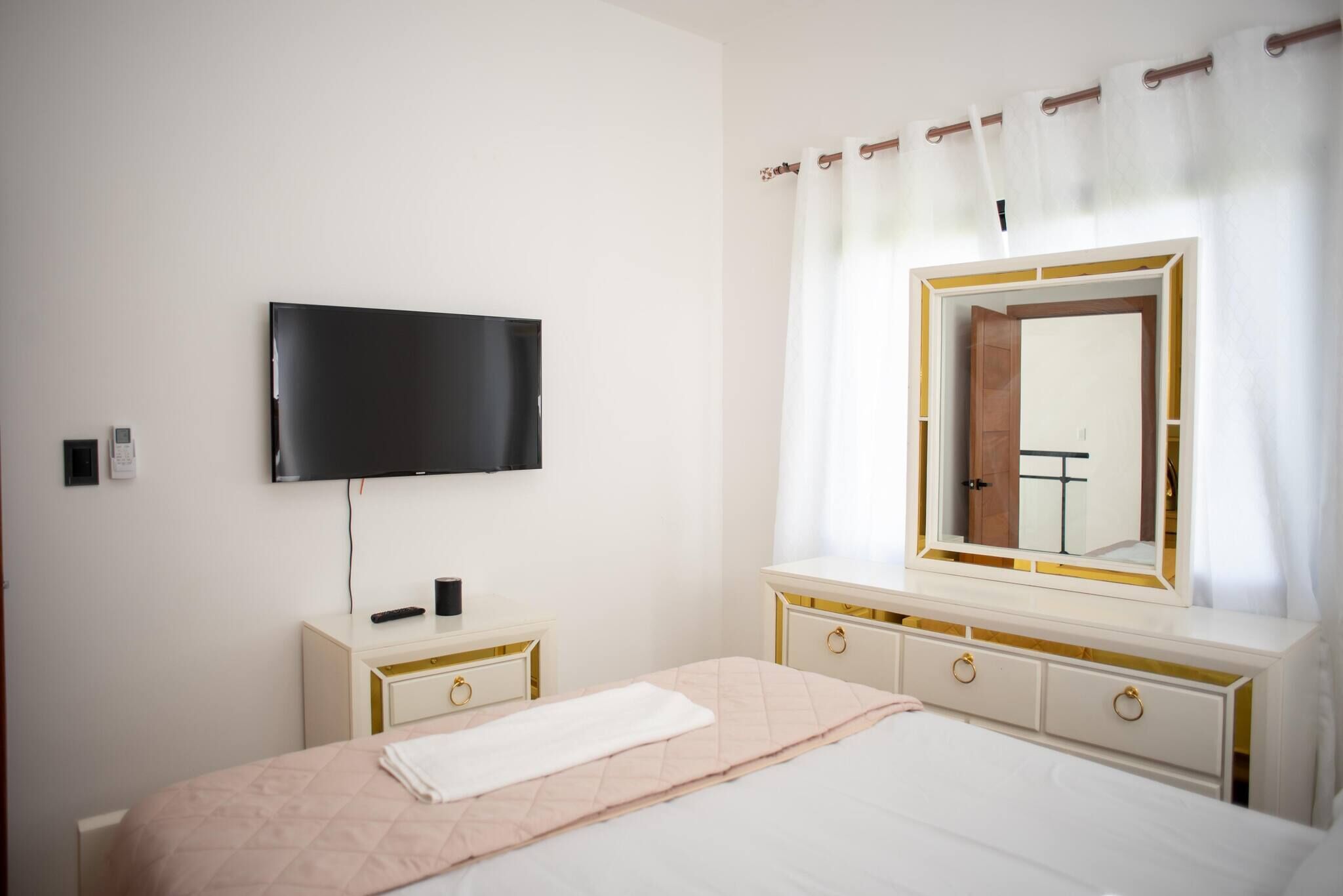 Une chambre à coucher comprenant un lit, un téléviseur fixé au mur, une commode et un miroir avec un rideau.