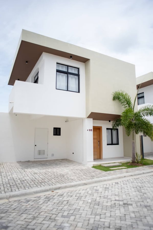 A modern two-story building with a flat roof, a single entrance with a wooden door, and a small garden area with a palm tree. - Luxurious Punta Cana House 3 Bedrooms, Shared Pool, BBQ (Punta Cana)