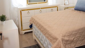 A bedroom with a bed, a bedside table, a dresser, a mirror, an air conditioning unit, and a window with curtains.