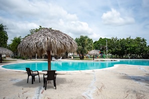 A swimming pool with a thatched umbrella, lounge chairs, and palm trees.