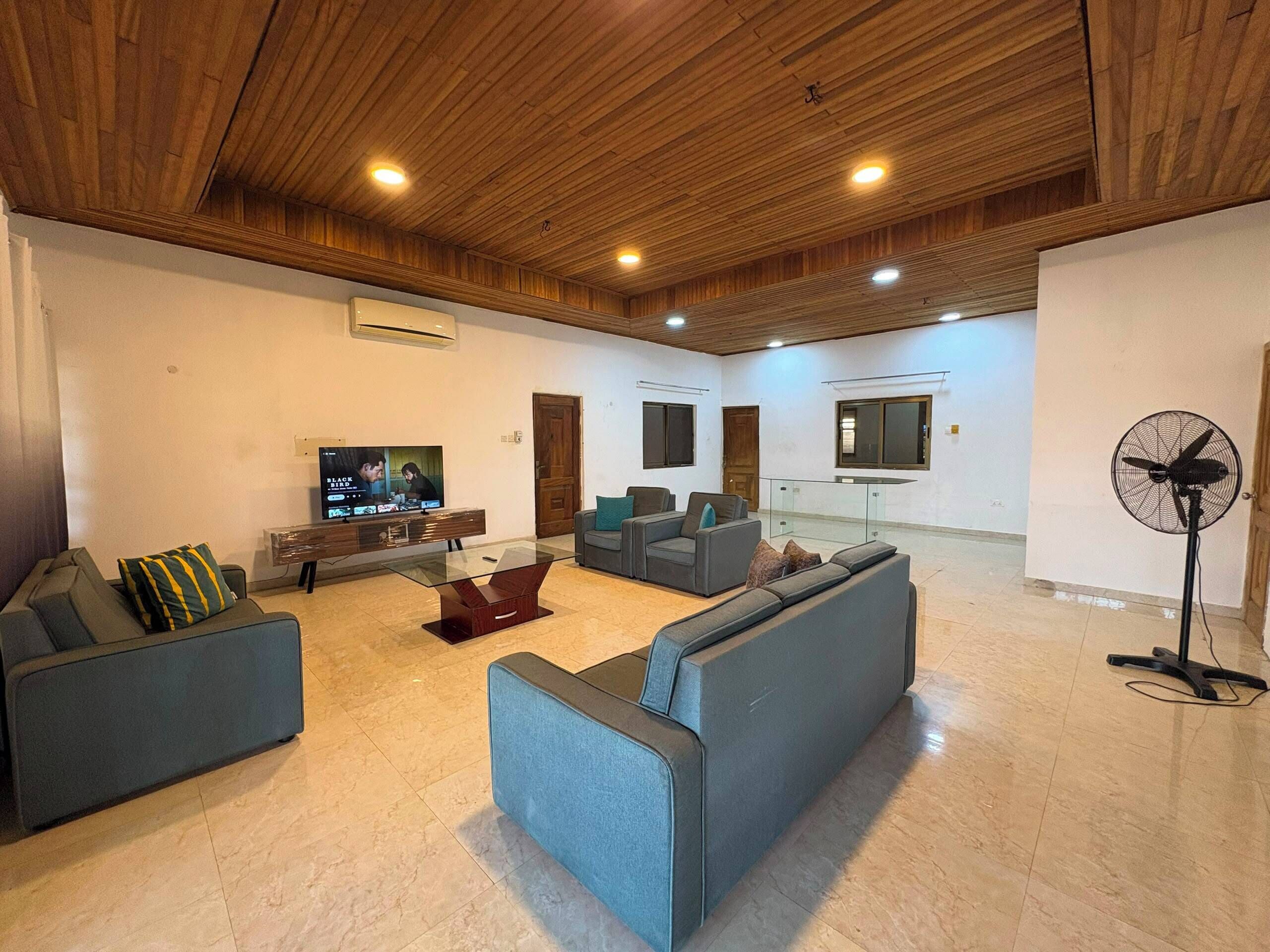 Amplia sala de estar con techo de madera, televisor de pantalla plana, varios sofás, una mesa de centro de vidrio y un ventilador de pie.
