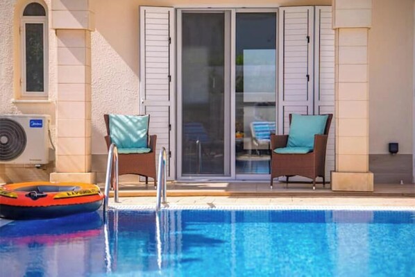 Una piscina con tobogán, dos sillas de descanso con cojines y un ventanal que da a una zona cubierta.