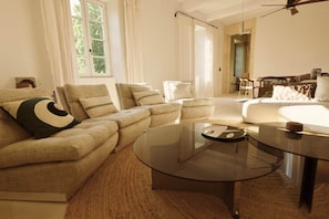 A living room with a sofa, armchairs, a glass coffee table, and a dining area in the background.
