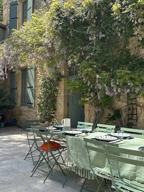 An outdoor dining area with tables and chairs, a stone building, and a tree with purple flowers.