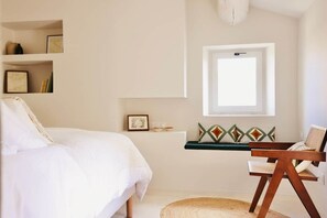 A bedroom with a bed, a chair, a shelf with decorative items, a window, and a ceiling light fixture.