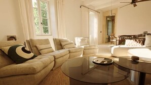 Een woonkamer met een bank, fauteuils, een glazen salontafel en een eethoek op de achtergrond.