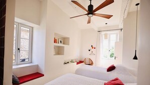 Une chambre avec deux lits, un ventilateur de plafond, une fenêtre avec un banc rouge et une étagère fixée au mur.