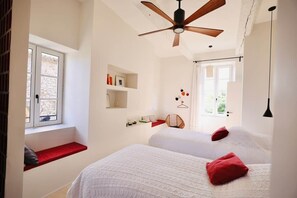 Une chambre avec deux lits, un ventilateur de plafond, une fenêtre avec un banc rouge et une étagère fixée au mur.