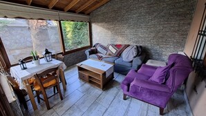 Una habitación acogedora con techo de madera, pared de piedra, un sofá púrpura, un sofá azul, una mesa de madera con sillas y una ventana con vistas a la vegetación.