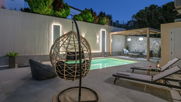 A poolside area with lounge chairs, a hanging chair, and a stone wall. - Luxury Villa Dimitra With Private Swimming Pool (Hersonissos)