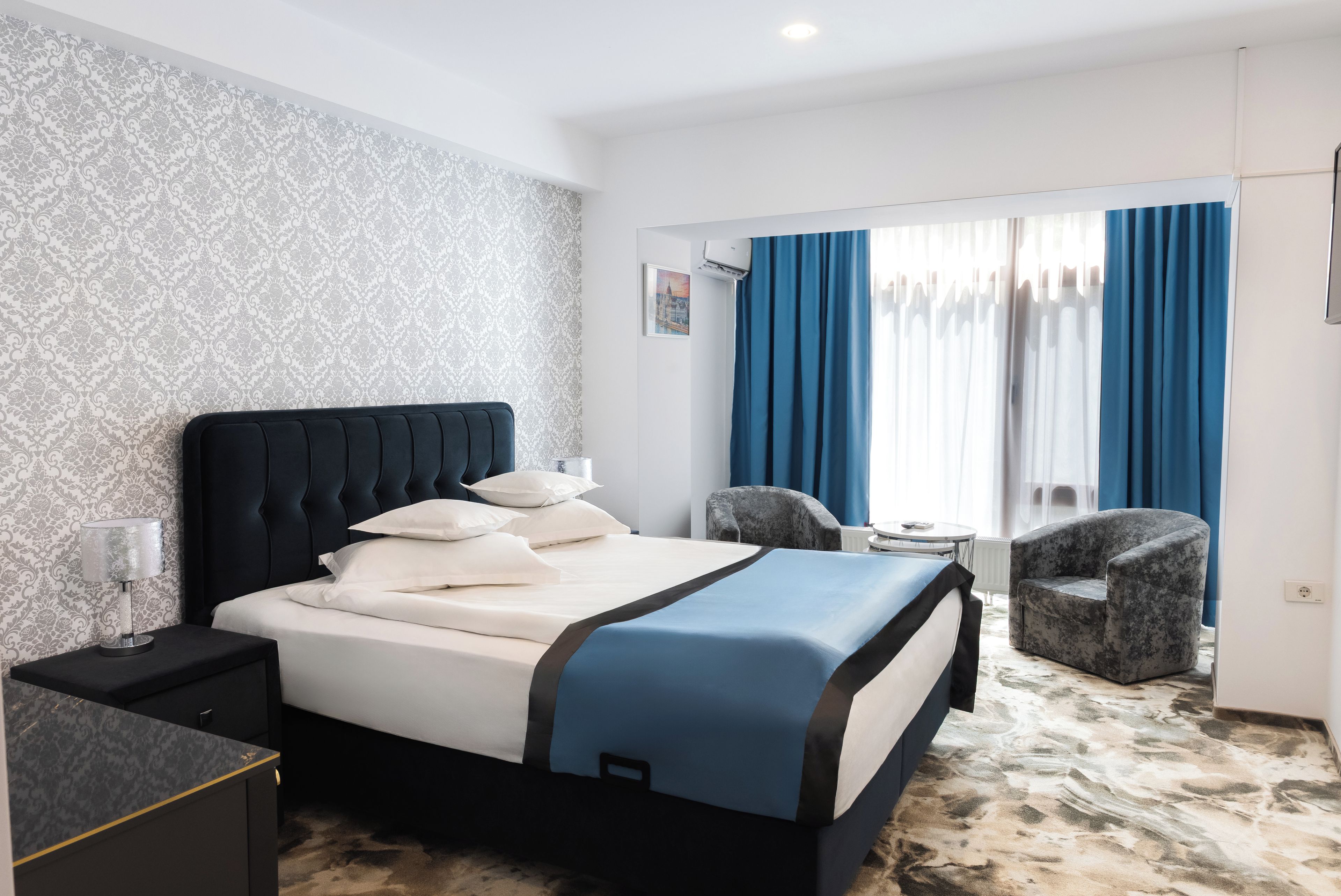Ein Hotelzimmer mit Bett, Nachttisch, Stuhl, kleinem Tisch und einem Fenster mit Vorhängen.