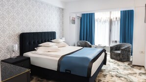 Una habitación de hotel con cama, mesita de noche, silla, una pequeña mesa y ventana con cortinas.