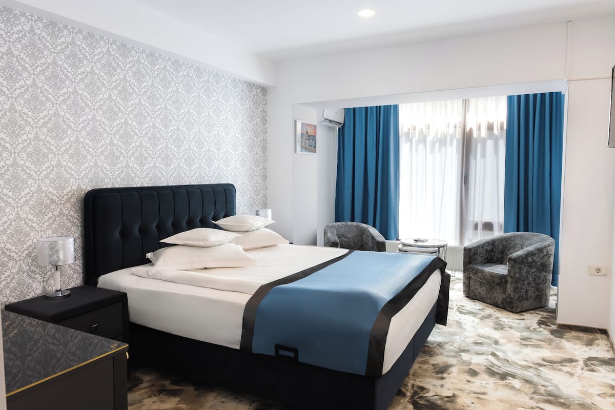 Ein Hotelzimmer mit Bett, Nachttisch, Stuhl, kleinem Tisch und einem Fenster mit Vorhängen.