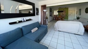 A modern room with a blue sofa, a bed, a dining area, and a kitchen in the background.
