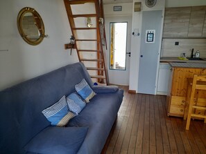 A room with a blue sofa, wooden ladder, and a kitchen area with wooden cabinets. - Apartment, sea view, pets welcome - 4 guests (Noirmoutier-en-l'Île)