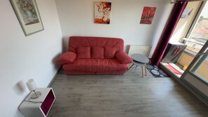 A red sofa, a small table, and a radiator in a room with a window. - La Corniche Large Studio (Courseulles-sur-Mer)