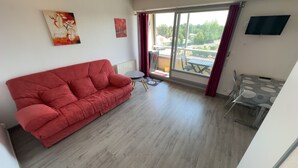 A red sofa, a small round table, a glass chair, a TV mounted on the wall, and a view of the outside through a sliding door. - La Corniche Large Studio (Courseulles-sur-Mer)