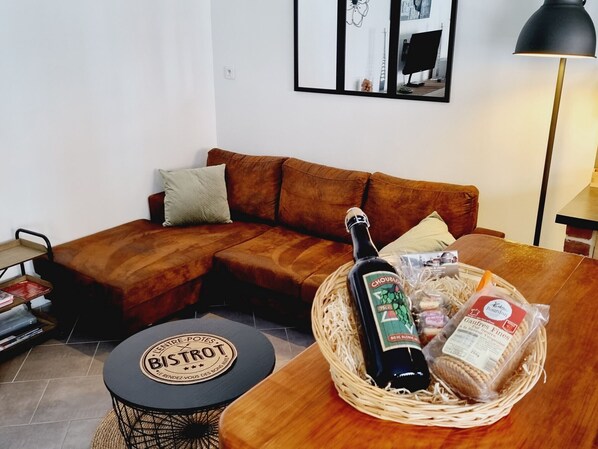 A living room with a brown sofa, a small round table with a logo, a bottle of wine, and a basket of snacks.