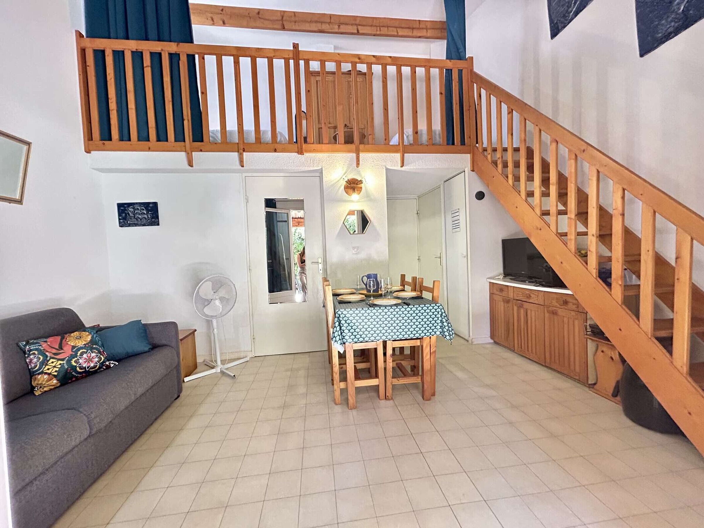 Ein Zimmer mit einer Holztreppe, einem Esstisch mit Stühlen, einem Sofa, einem Ventilator und einem Fernseher.