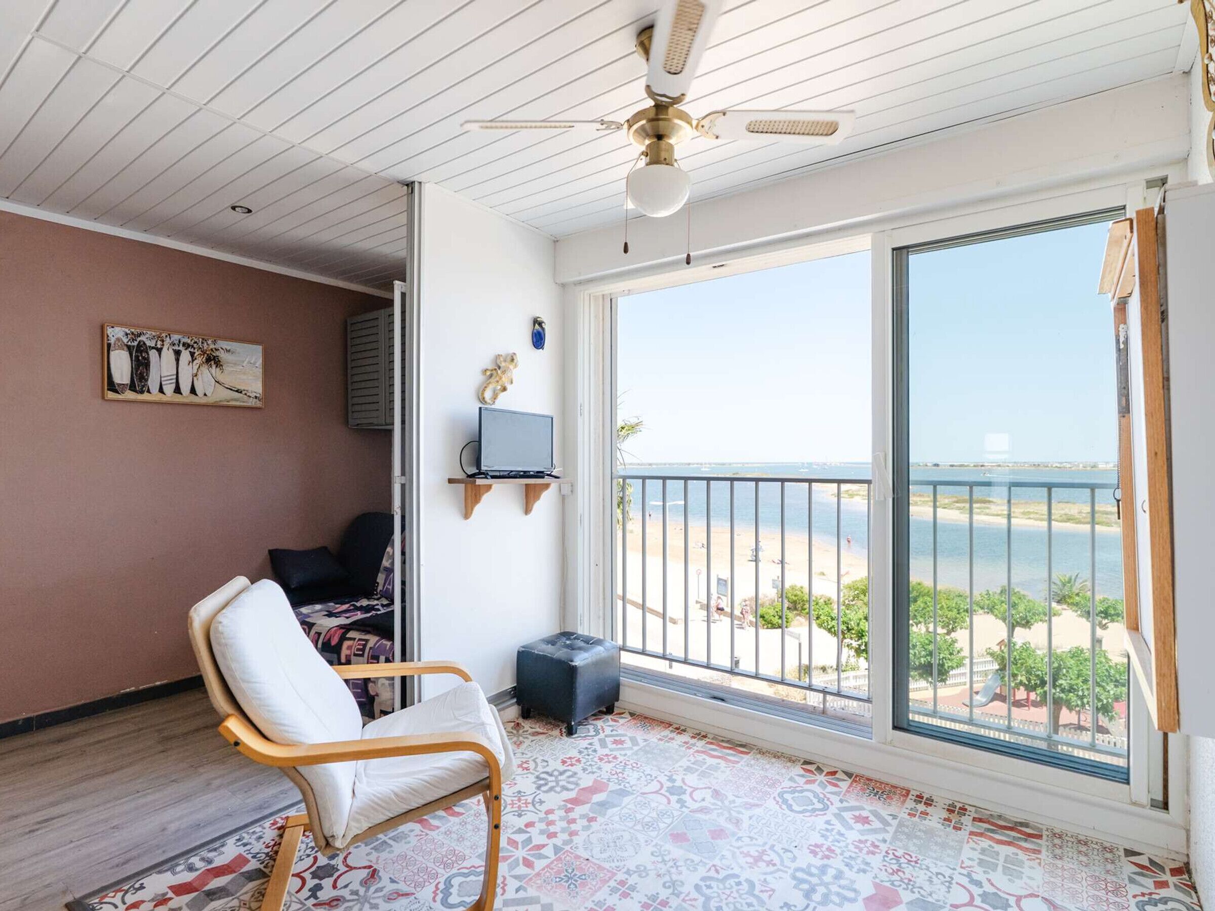 Une pièce dotée d’une grande porte-fenêtre coulissante donnant sur un balcon, d’un ventilateur de plafond, d’un téléviseur posé sur une étagère, d’un tapis à motifs et d’un fauteuil.