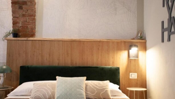 A hotel room with a wooden headboard, a bed with patterned bedding, a small table, and a wall-mounted light fixture. - Switch holiday 314 (La Spezia)