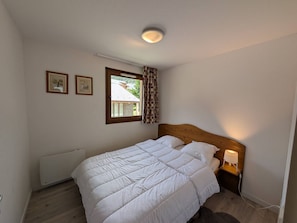 Een slaapkamer met een houten hoofdeinde, een witte dekbedovertrek, een radiator, een raam met gordijnen en een wandlamp.